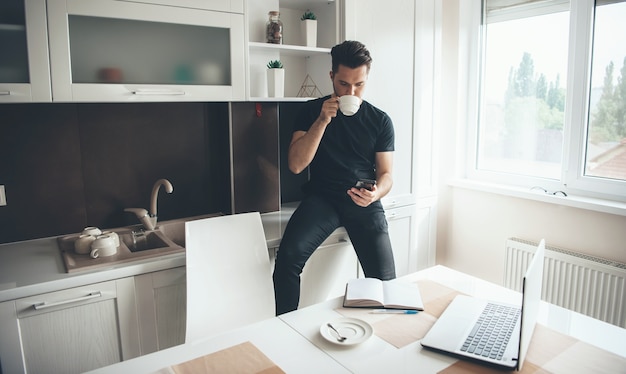 Joven empresario caucásico que trabaja desde casa usando el teléfono y la computadora portátil con el libro está tomando un café mientras hace una pausa en la cocina