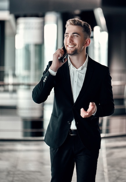 Un joven empresario en un centro de negocios con un teléfono con un traje negro y camisa blanca sonriendo y hablando por teléfono