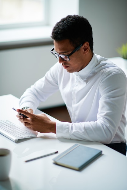 Joven empresario contemporáneo en anteojos y camisa blanca sentado junto al escritorio y desplazándose en el teléfono inteligente mientras planifica el trabajo en la oficina