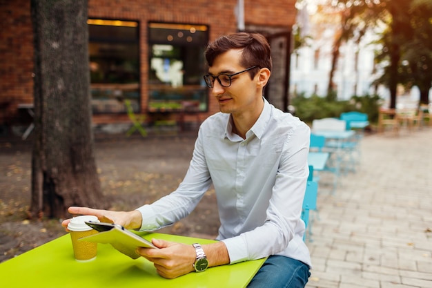 Joven empresario disfruta de un café en la cafetería al aire libre con tableta. Chico guapo bebiendo té en la calle de la ciudad con tableta