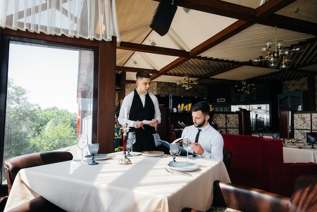 Un joven empresario en un elegante restaurante examina el menú y hace un pedido a un joven camarero con un elegante delantal. Servicio al Cliente.