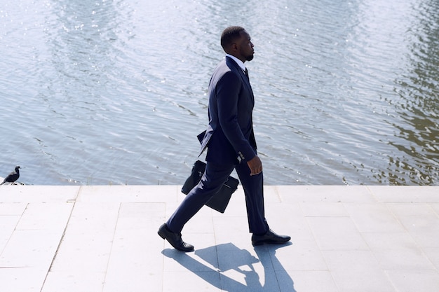 Joven empresario elegante en traje azul oscuro moviéndose por la acera frente a la cámara a lo largo de la orilla del río mientras se apresura a trabajar por la mañana