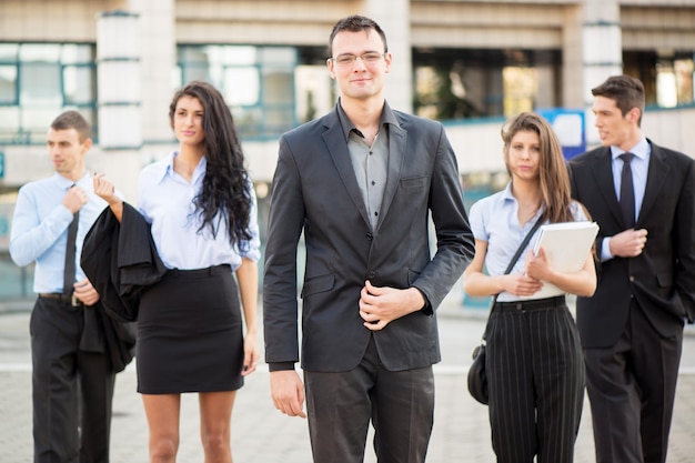 Joven empresario, elegantemente vestido, orgullosamente con su equipo de jóvenes empresarias y empresarios frente al edificio de oficinas.