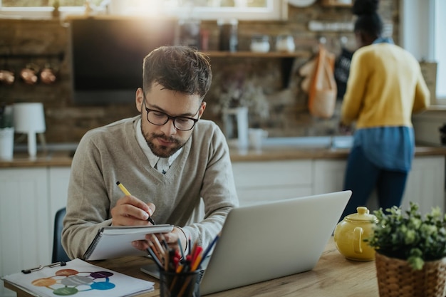 Joven empresario escribiendo notas mientras trabaja en una computadora en casa