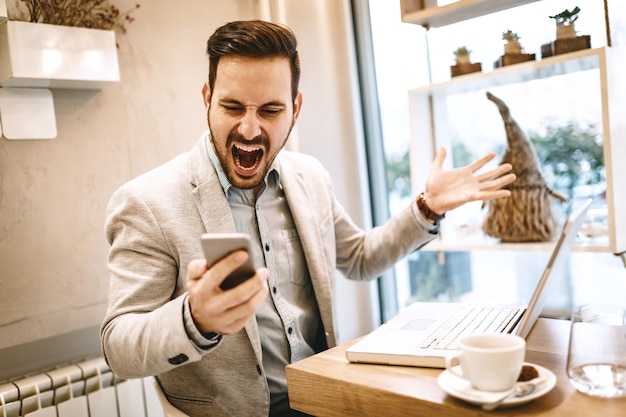 Joven empresario estresado gritando en el teléfono inteligente y trabajando en la computadora portátil en el café.