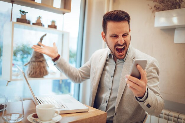 Joven empresario estresado gritando en el teléfono inteligente y trabajando en la computadora portátil en el café.