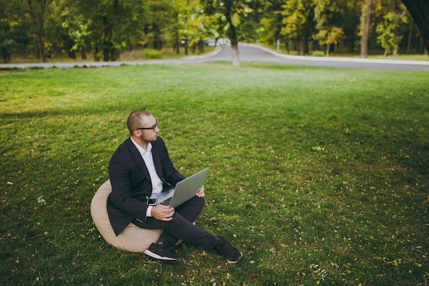 Joven empresario de éxito en camisa blanca, traje clásico, gafas. El hombre se sienta en un puf suave, trabajando en un ordenador portátil en el parque de la ciudad en césped verde al aire libre en la naturaleza. Oficina móvil, concepto de negocio.