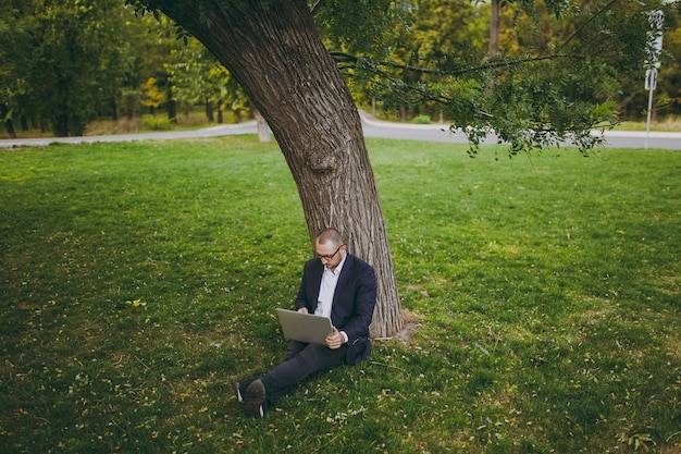 Joven empresario de éxito en camisa blanca, traje clásico, gafas. El hombre se sienta en el suelo de la hierba, trabaja en la computadora portátil de la PC en el parque de la ciudad en césped verde al aire libre en la naturaleza. Oficina móvil, concepto de negocio.