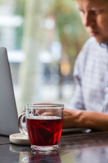 Joven empresario de éxito sentado en un café, bebiendo té rojo saludable y trabajando en una computadora portátil. Composición de estilo de vida con luz natural.
