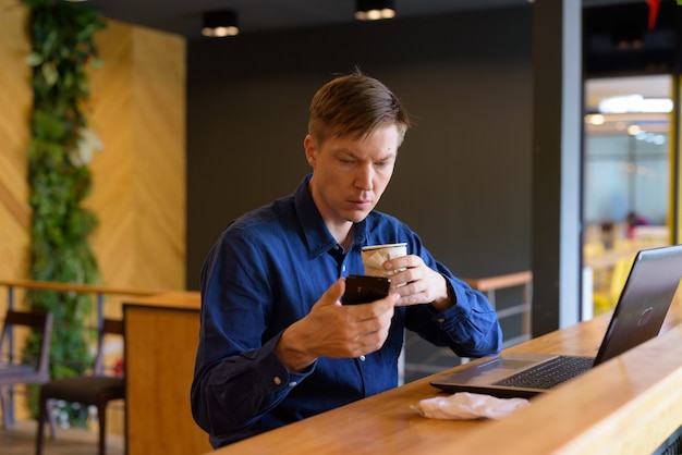 Joven empresario guapo con café usando teléfono y sentado con distancia en la cafetería.