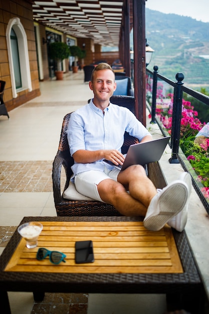 Joven empresario guapo con una camisa blanca y pantalones cortos se sienta con una computadora portátil en un café en la mesa. Trabaja descansando. Enfoque selectivo