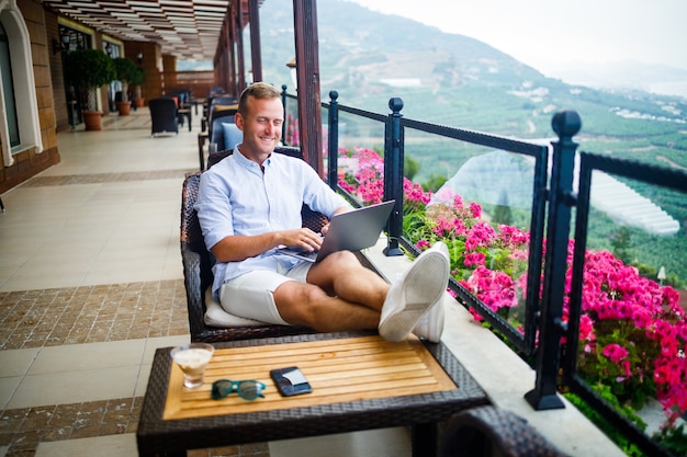 Joven empresario guapo con una camisa blanca y pantalones cortos se sienta con una computadora portátil en un café en la mesa. Trabajar descansando