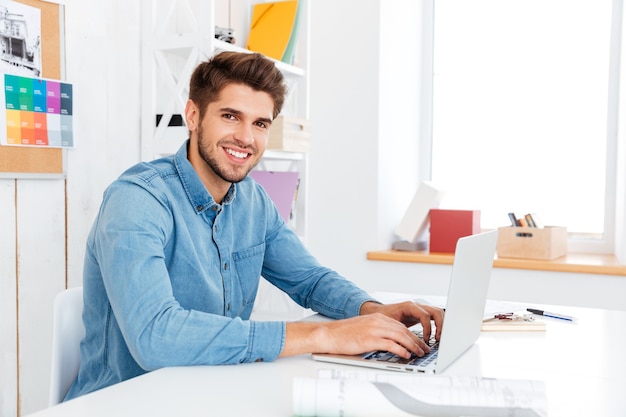 Joven empresario guapo escribiendo en la computadora portátil y mirando a la cámara mientras está sentado en la oficina