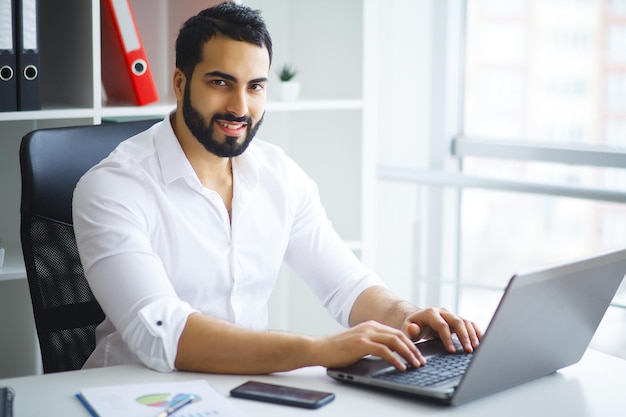 Joven empresario guapo sentado en el escritorio y usando la computadora portátil en la oficina.