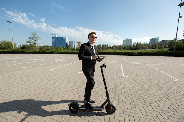 Joven empresario guapo en traje monta un scooter eléctrico y mira una computadora portátil.