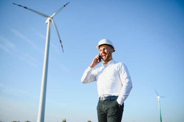 Joven empresario hablando con el gerente mediante teléfono inteligente en el campo de la turbina eólica, concepto de energía verde