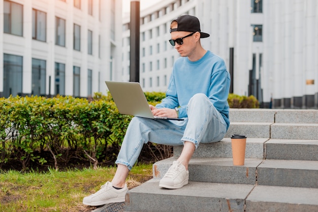 Joven empresario hipster barbudo se encuentra en la calle de la ciudad tiene una taza de café y usa una tableta. Edificio de cristal moderno hombre que trabaja blogs chateando en las redes sociales en línea