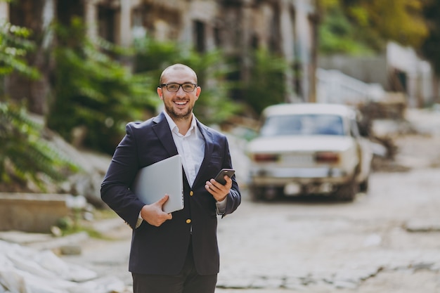 Joven empresario inteligente exitoso en camisa blanca, traje clásico, gafas. Hombre de pie con el teléfono de la computadora del ordenador portátil cerca de las ruinas, escombros, edificio de piedra al aire libre. Oficina móvil, negocio, concepto de trabajo.