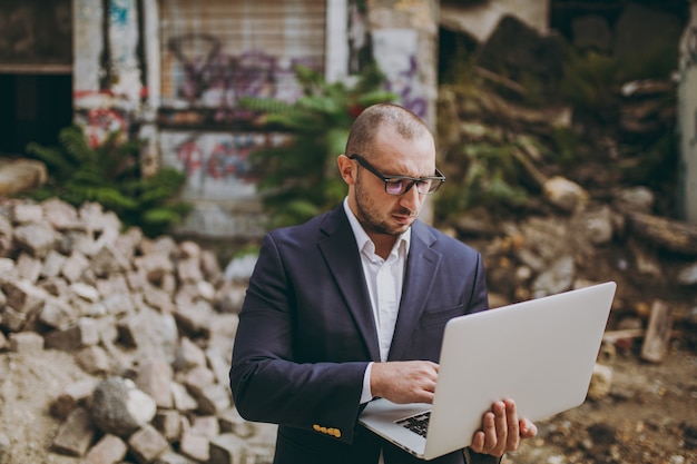 Joven empresario inteligente exitoso en camisa blanca, traje clásico, gafas. Hombre de pie y trabajando en una computadora portátil cerca de ruinas, escombros, edificio de piedra al aire libre. Oficina móvil, concepto de negocio.