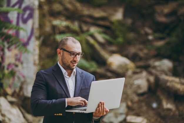 Joven empresario inteligente exitoso en camisa blanca, traje clásico, gafas. Hombre de pie y trabajando en una computadora portátil cerca de ruinas, escombros, edificio de piedra al aire libre. Oficina móvil, concepto de negocio.