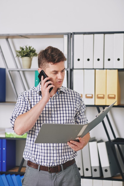 Joven empresario leyendo el documento en la carpeta abierta y hablando por teléfono con el cliente o compañero de trabajo