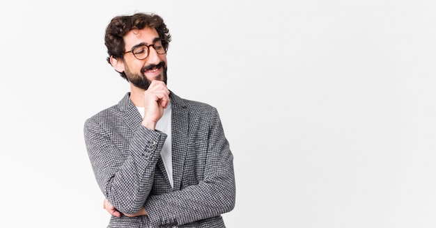 Joven empresario loco sonriendo con una expresión feliz y segura con la mano en la barbilla, preguntándose y mirando a un lado contra la pared plana