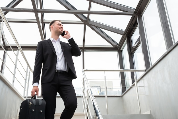 Joven empresario con maleta en elegante traje hablando por teléfono mientras camina por las escaleras en el moderno edificio empresarial