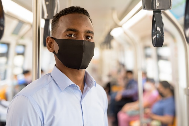 Joven empresario con máscara para protegerse del brote de virus corona de pie con distancia dentro del tren