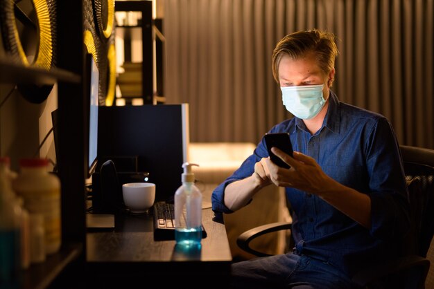 Joven empresario con máscara usando teléfono en casa mientras trabaja de noche