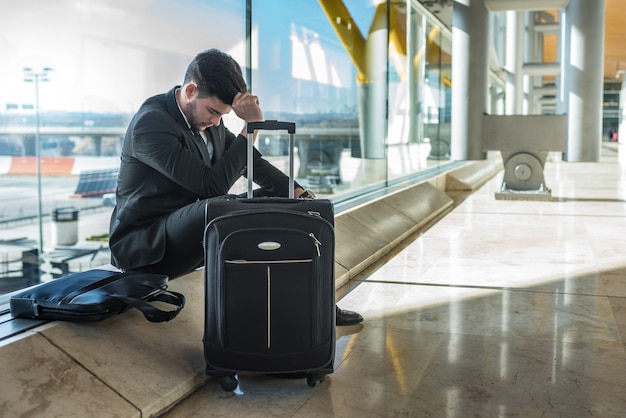 Joven empresario molesto en el aeropuerto esperando su vuelo retrasado con equipaje