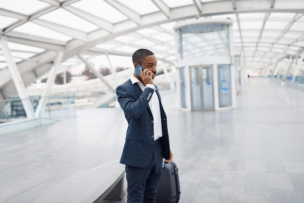 Joven empresario negro haciendo llamadas móviles mientras está de pie con equipaje en el aeropuerto