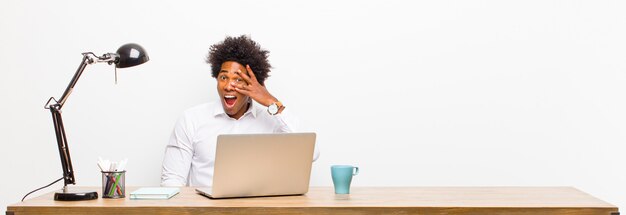 Joven empresario negro mirando sorprendido, asustado o aterrorizado, cubriéndose la cara con la mano y mirando entre los dedos sobre un escritorio