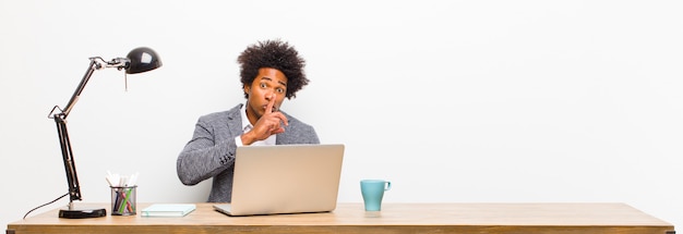 Joven empresario negro pidiendo silencio y silencio, gesticulando con el dedo frente a la boca, diciendo shh o guardando un secreto en un escritorio