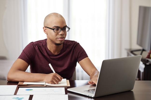 Joven empresario negro revisando correos electrónicos y documentos financieros en una computadora portátil y tomando notas en el planificador