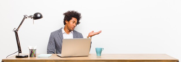 Joven empresario negro sintiéndose feliz y sonriendo casualmente, mirando a un objeto o concepto sostenido en la mano al lado de un escritorio