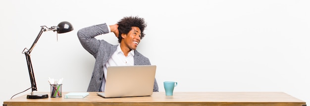 Joven empresario negro sonriendo alegremente y casualmente, tomando mano a cabeza con una mirada positiva, feliz y segura en un escritorio