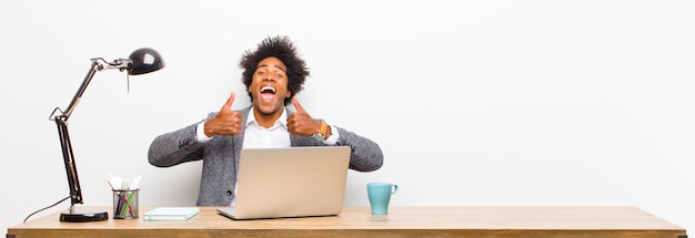 Joven empresario negro sonriendo ampliamente mirando feliz, positivo, seguro y exitoso, con ambos pulgares en un escritorio