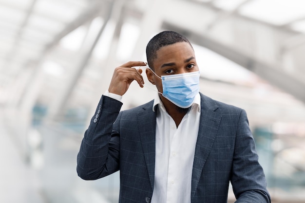 Joven empresario negro tomando mascarilla médica mientras camina en el aeropuerto