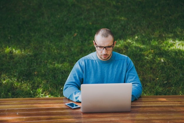 Joven empresario o estudiante exitoso hombre inteligente en gafas de camisa azul casual sentado a la mesa con el teléfono móvil en el parque de la ciudad usando laptop trabajando al aire libre sobre fondo verde. Concepto de oficina móvil.