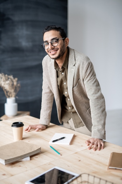 Joven empresario o gerente sonriente en ropa formal que le mira mientras se inclina sobre la mesa de madera durante el trabajo