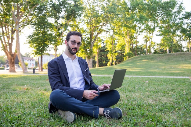 Joven empresario en el parque responde un correo electrónico mientras está sentado en el césped Trabaja en cualquier lugar