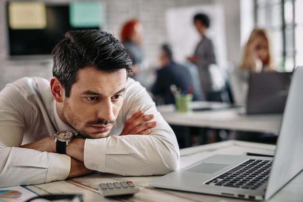 Joven empresario pensando en algo mientras se apoya en su escritorio y usa la computadora en el trabajo Hay gente en el fondo