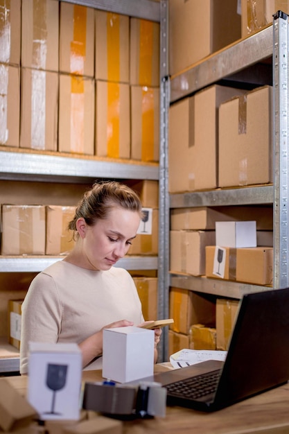 Joven empresario de pequeña empresa que comprueba los pedidos de Internet en su teléfono inteligente y prepara artículos en cajas para su entrega a los clientes