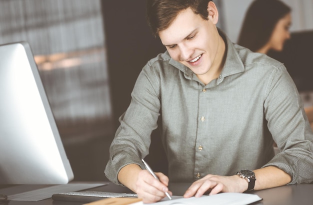 Un joven empresario y programador con un pantalones verde está escribiendo algunas notas en su plan de negocios, mientras está sentado en el escritorio de un gabinete junto con su colega en el fondo. Negocios