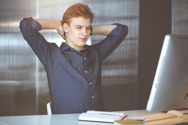 Joven empresario y programador rubio estirando las manos después de trabajar duro con la computadora en una oficina oscura, resplandor de luz en el fondo.