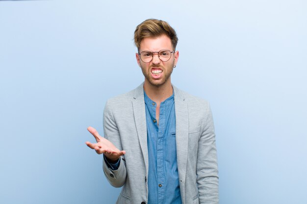 Joven empresario que parece enojado, molesto y frustrado gritando wtf o qué te pasa contra la pared azul