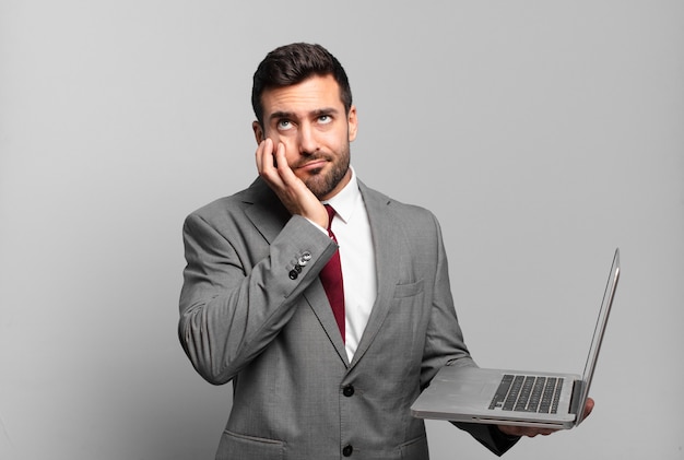 Joven empresario que se siente aburrido, frustrado y con sueño después de una tarea tediosa, aburrida y tediosa, sosteniendo la cara con la mano y sosteniendo una computadora portátil