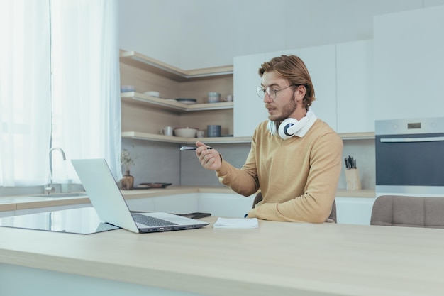 Joven empresario que trabaja de forma remota desde casa sentado en la cocina usando una computadora portátil para videollamadas