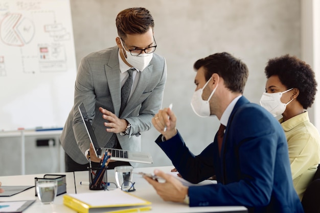 Joven empresario que usa laptop mientras habla con sus compañeros de trabajo durante una reunión de negocios en la oficina. Llevan máscaras faciales protectoras debido a la epidemia de coronavirus.