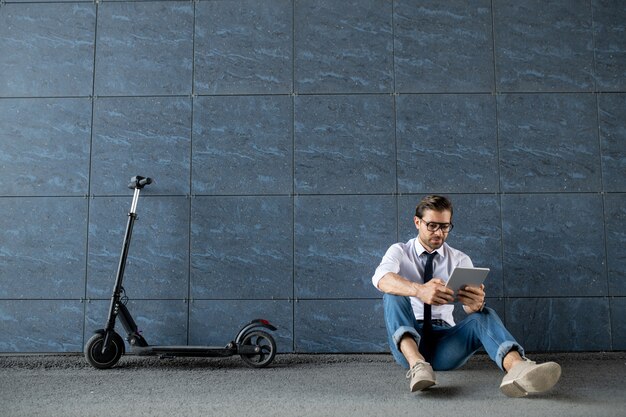 Joven empresario relajado en ropa casual elegante sentado junto a la pared del edificio y desplazamiento en tableta digital